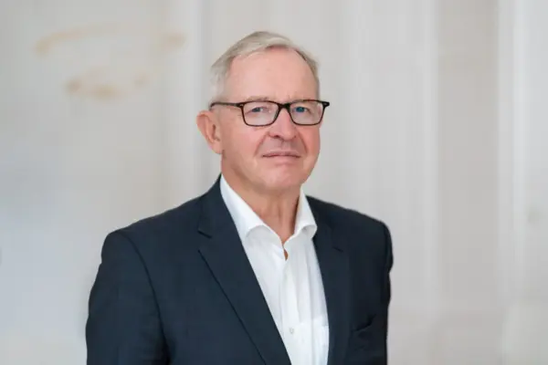 Prof. Dr. Joachim Erdmann Porträtfoto von Prof. Dr. Joachim Erdmann: älterer Mann mit kurzen weißgrauen Haaren und dunkler eckiger Brille, blickt ruhig in die Kamera, trägt einen dunkelblauen Blazer mit weißem Hemd ohne Krawatte, heller eleganter Hintergrund.