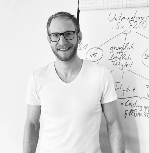 Alt-Text-Vorschlag: „Porträtfoto von Christoph Glüsenkamp: Mann mit kurzen Haaren, Bart und markanter schwarzer Brille, lächelt in die Kamera, trägt ein weißes T-Shirt, steht vor einem Flipchart mit handgeschriebenen Notizen zu rechtlichen Fachbegriffen, Schwarz-Weiß-Aufnahme.