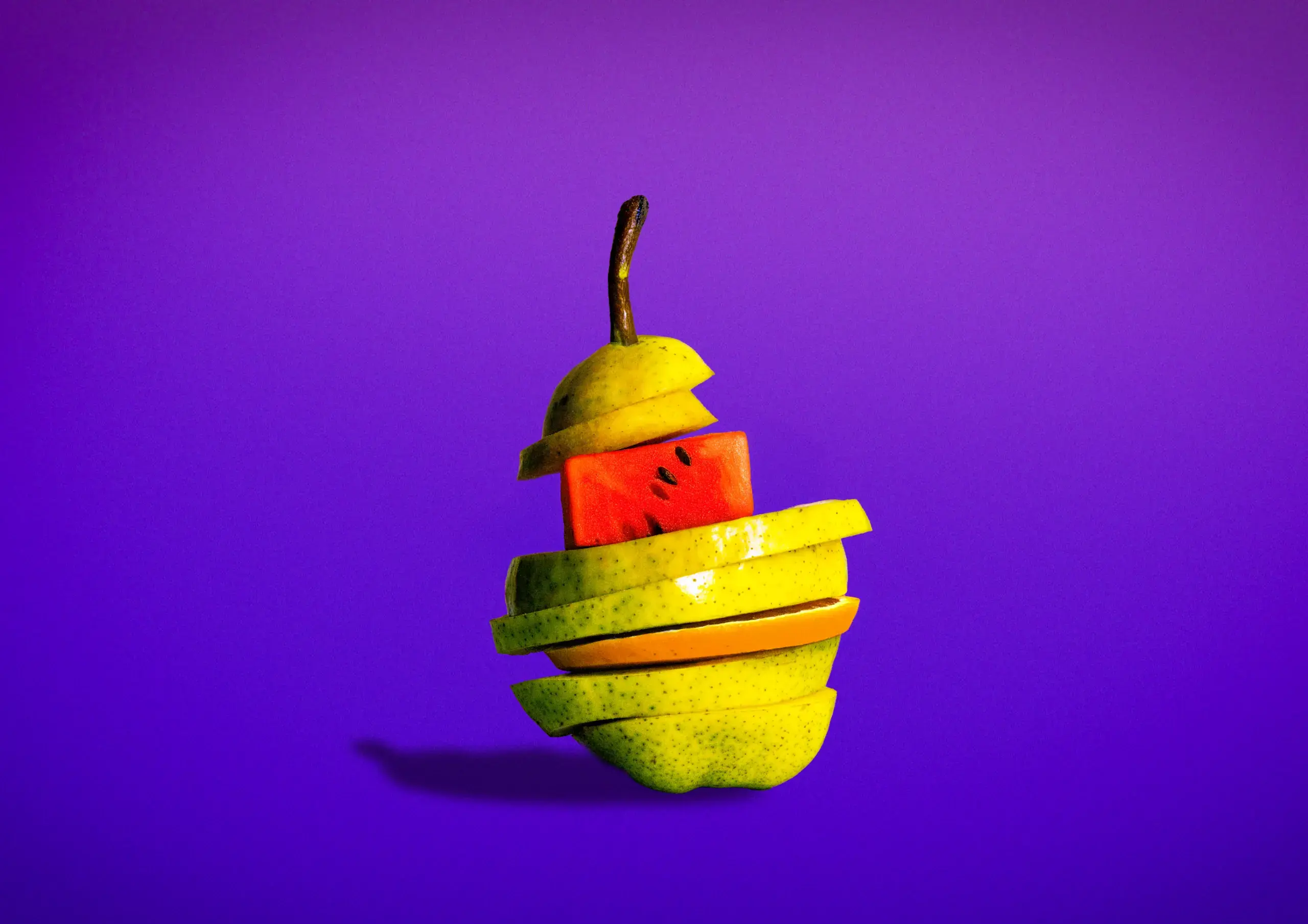 Kreatives Stillleben: Eine in horizontale Scheiben geschnittene Birne, in die verschiedene Fruchtscheiben – darunter Wassermelone und Orange – eingeschoben sind, schwebt vor einem leuchtend violetten Hintergrund.
