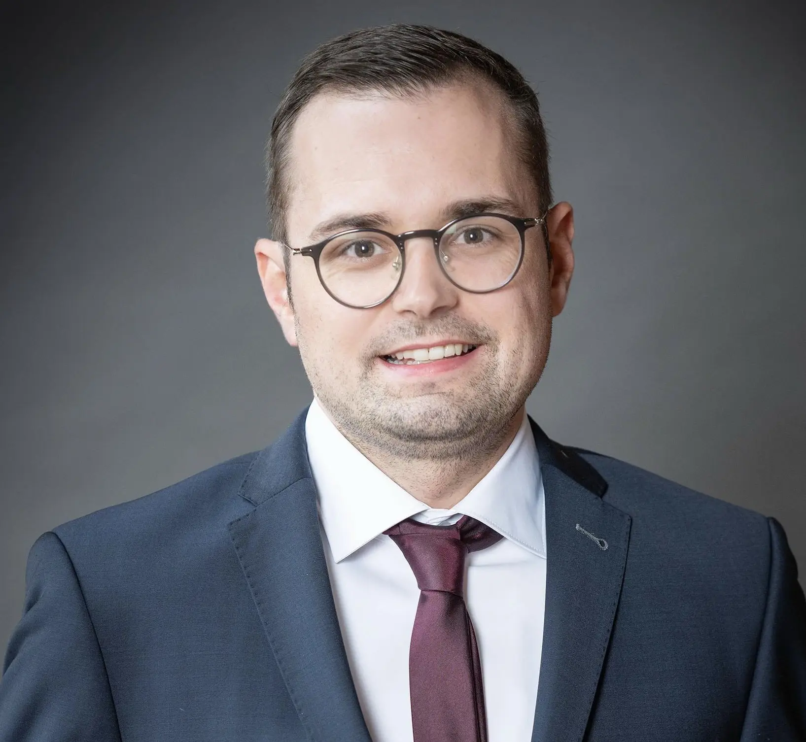 Julius Rega Porträtfoto eines lächelnden Mannes mit Brille, kurzen dunklen Haaren und leichtem Bartschatten, in dunkelblauem Anzug, weißem Hemd und weinroter Krawatte, vor grauem Hintergrund.